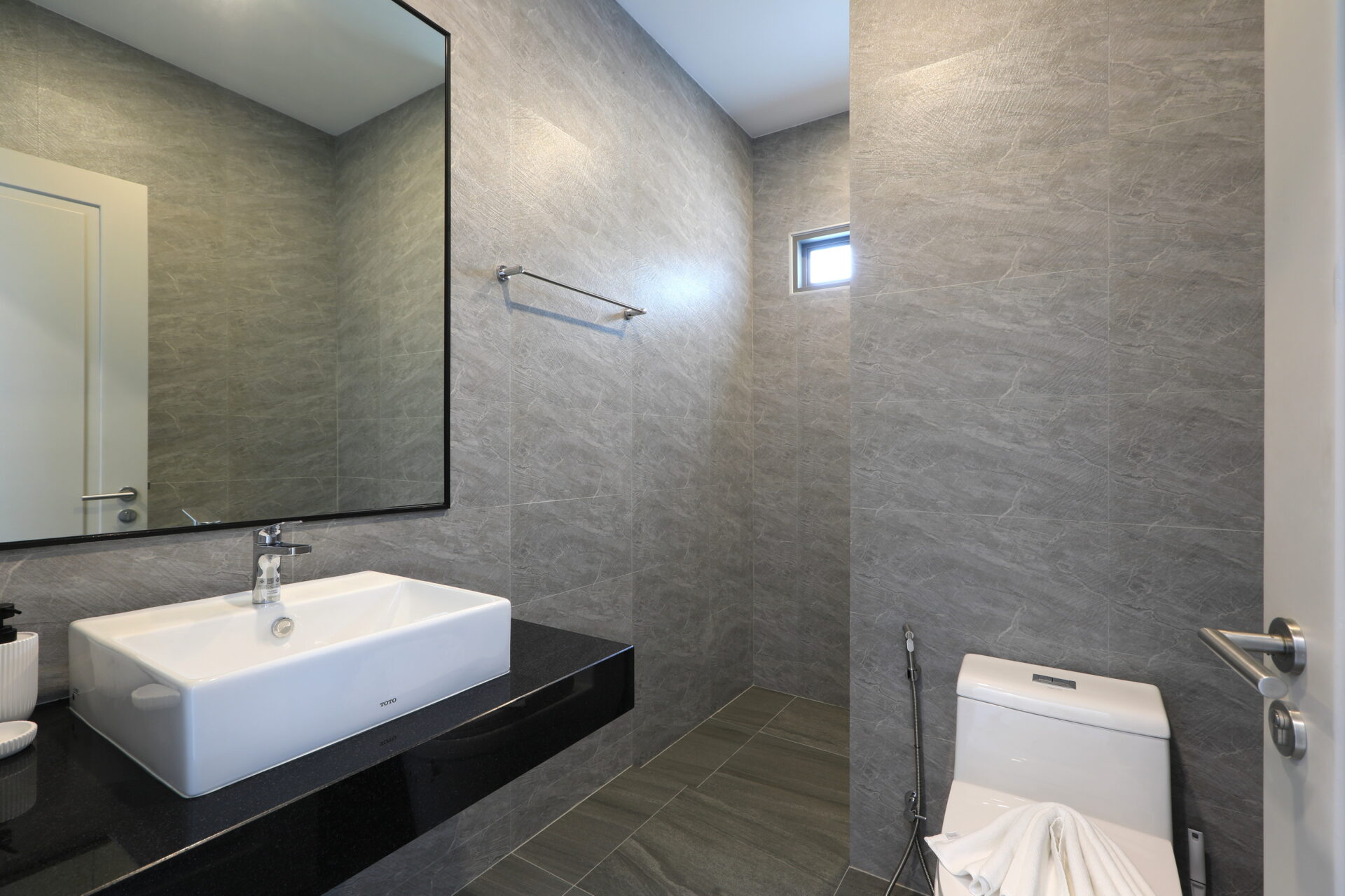 Modern grey-tiled bathroom with a white rectangular sink on a black countertop, large wall mirror, towel bar, and a white toilet.