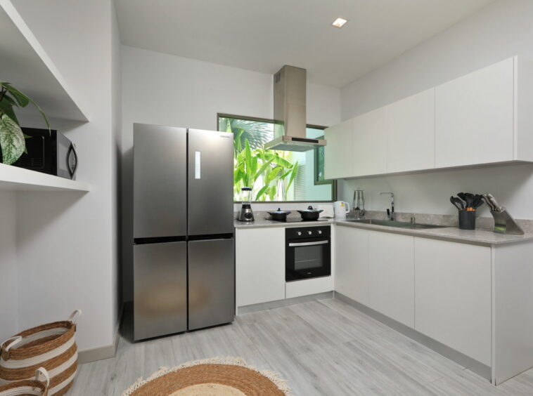 Modern white kitchen with a stainless steel Samsung refrigerator, built-in oven, and gray countertops; green plants visible through the window.