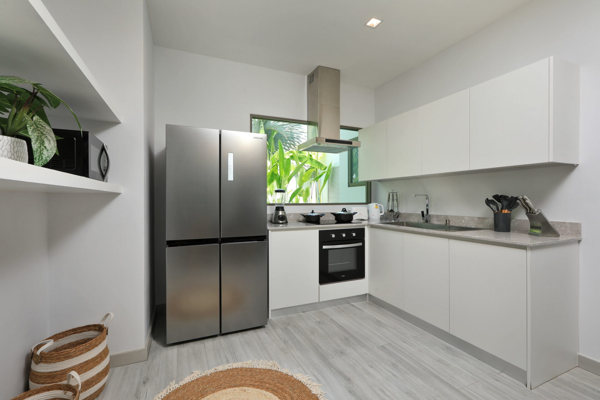 Modern white kitchen with a stainless steel Samsung refrigerator, built-in oven, and gray countertops; green plants visible through the window.