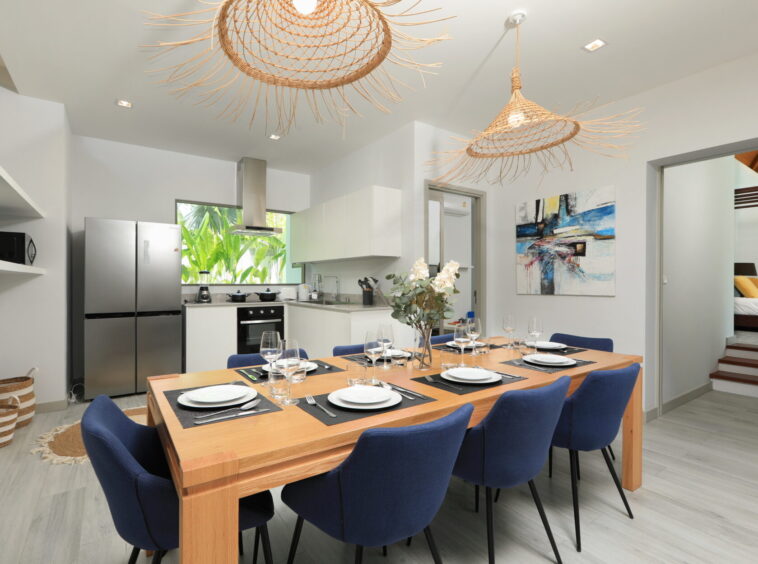 Open-plan dining room with a long wooden table set for twelve, blue upholstered chairs, and a modern kitchen in the background.