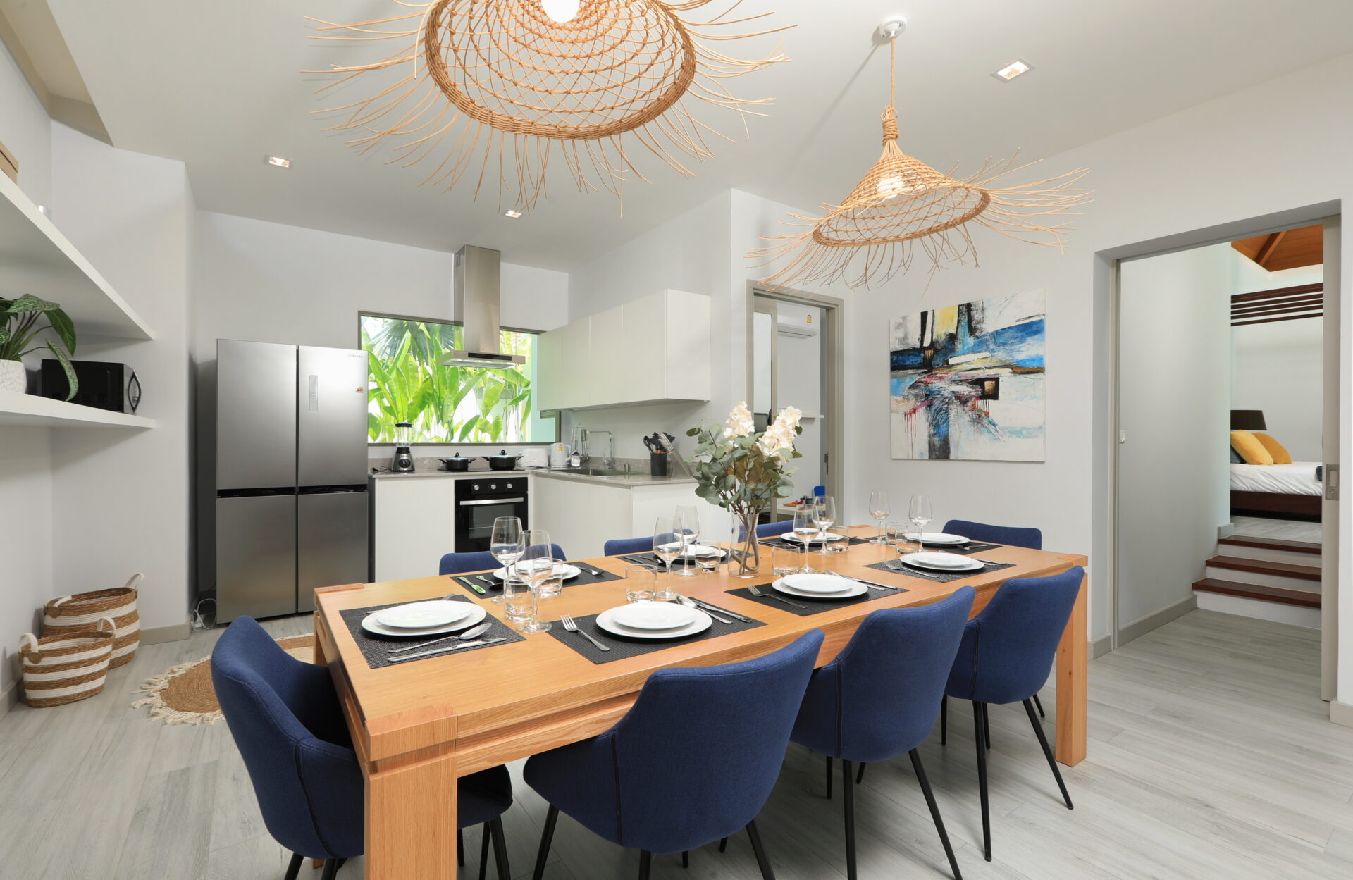 Open-plan dining room with a long wooden table set for twelve, blue upholstered chairs, and a modern kitchen in the background.