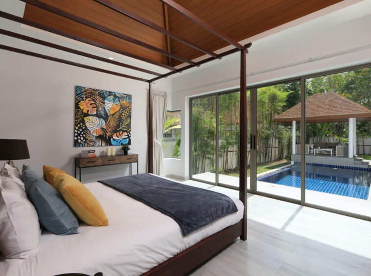 Wood framed canopy bed in a bright bedroom with colorful throw pillows and a view of a pool outside through large glass doors.