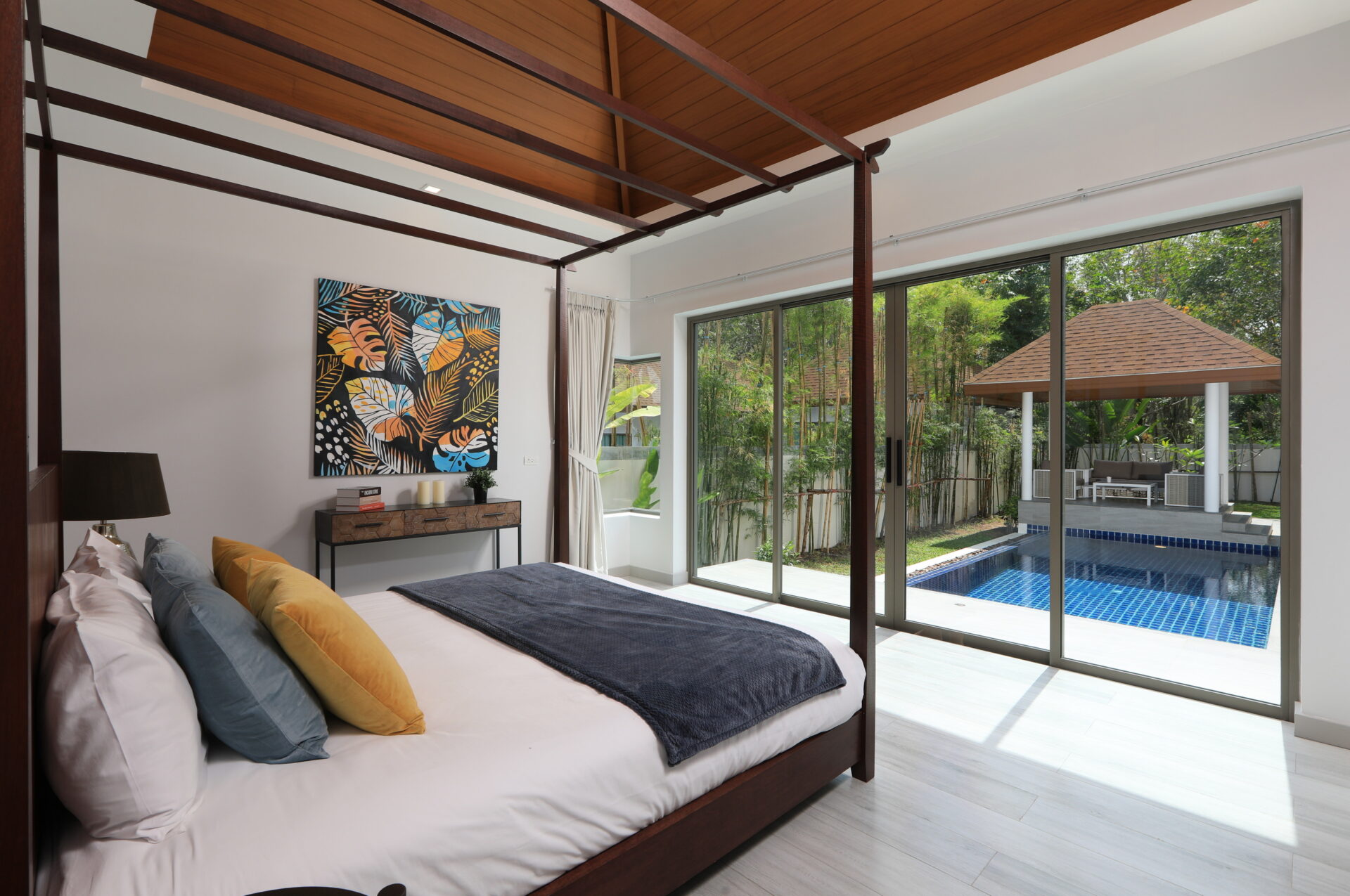 Wood framed canopy bed in a bright bedroom with colorful throw pillows and a view of a pool outside through large glass doors.