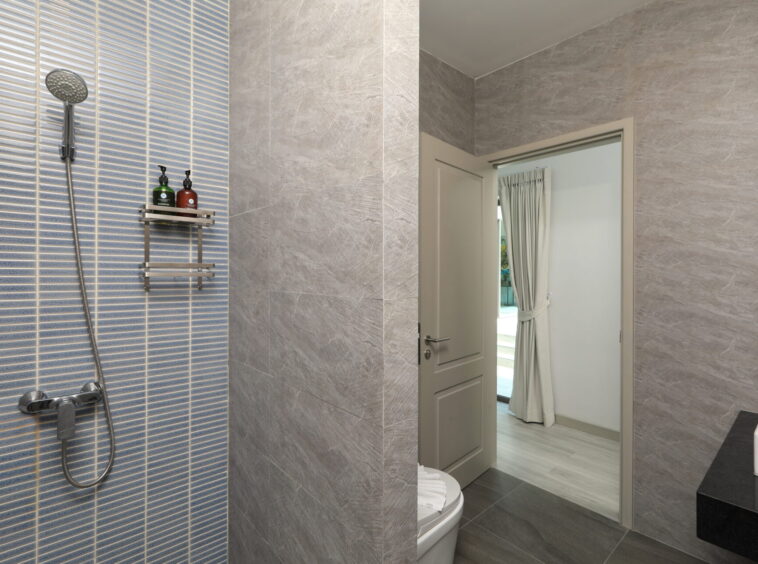 Modern bathroom featuring a blue-tiled shower wall with a handheld showerhead and metal shelf holding soap bottles; grey stone walls and a doorway to an adjacent room with a window and curtains.