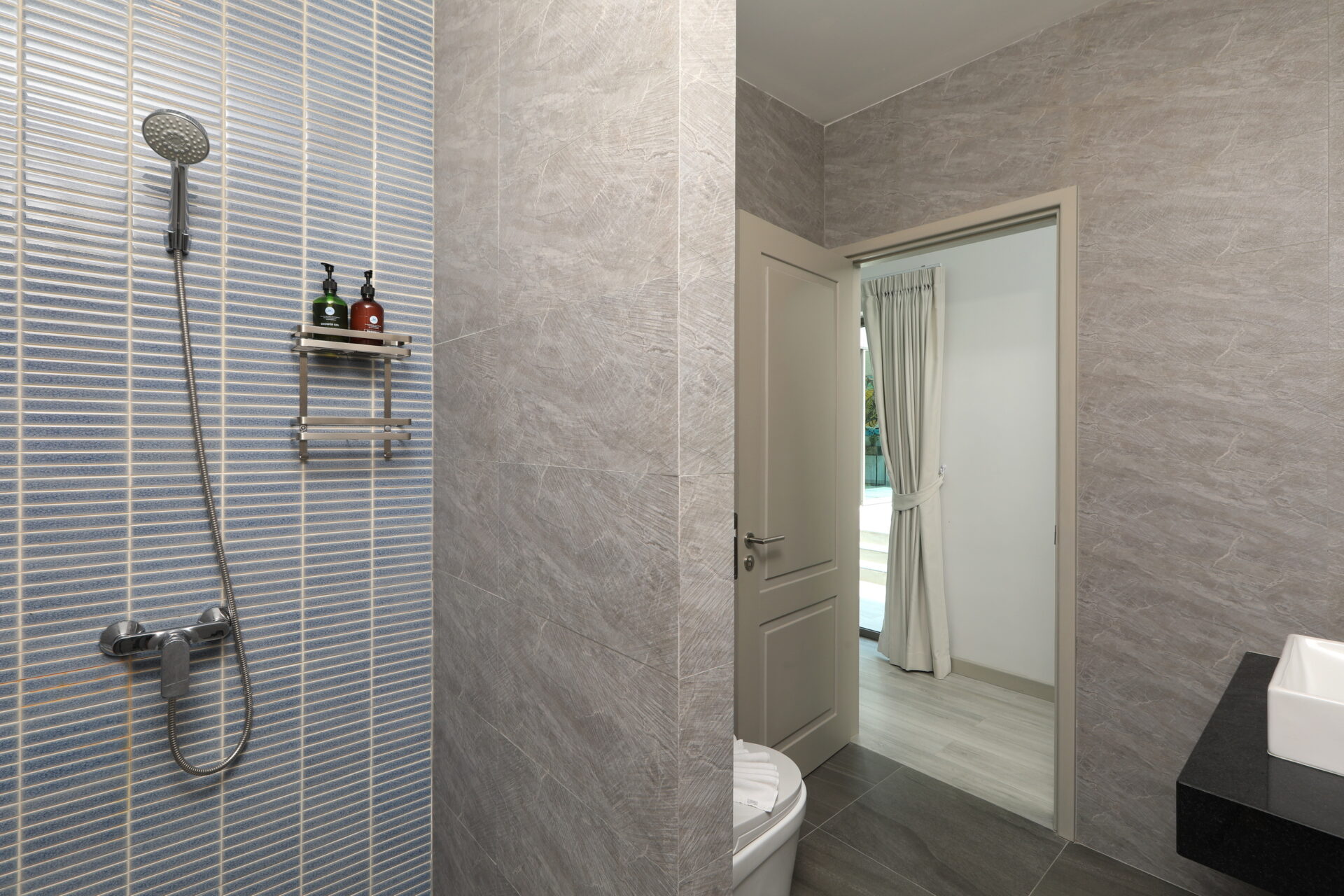 Modern bathroom featuring a blue-tiled shower wall with a handheld showerhead and metal shelf holding soap bottles; grey stone walls and a doorway to an adjacent room with a window and curtains.