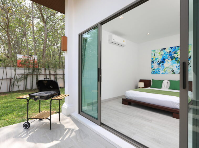 Bedroom visible through open sliding glass doors to a patio with a charcoal grill, wooden table, and green lawn outside; inside a white room with a king bed, green accents, and abstract blue-green artwork on the wall
