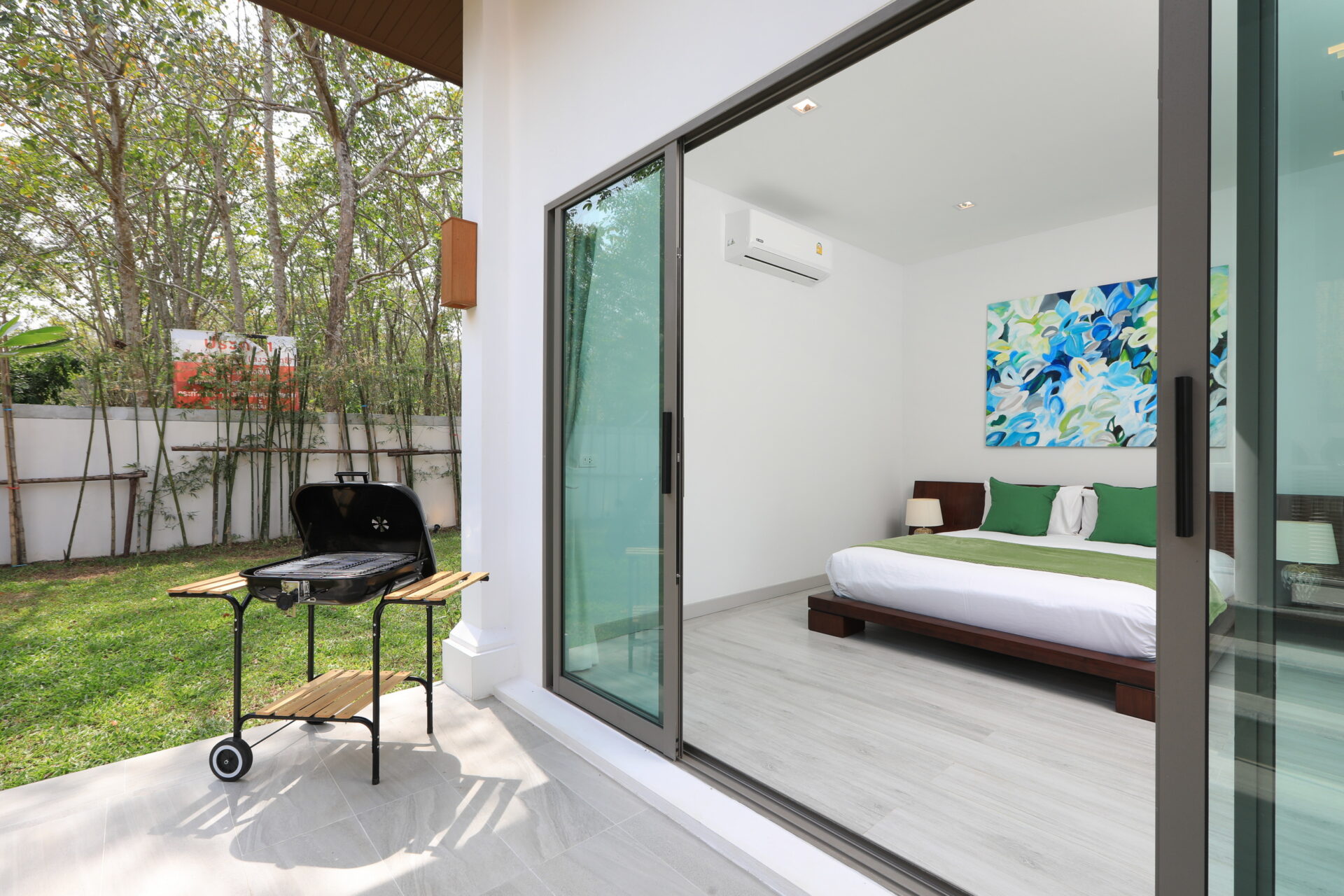 Bedroom visible through open sliding glass doors to a patio with a charcoal grill, wooden table, and green lawn outside; inside a white room with a king bed, green accents, and abstract blue-green artwork on the wall