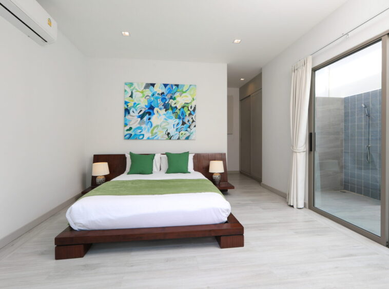 Modern bedroom with a low wooden platform bed, white linens, green pillows, a colorful abstract painting, and a glass door opening to an outdoor shower area.