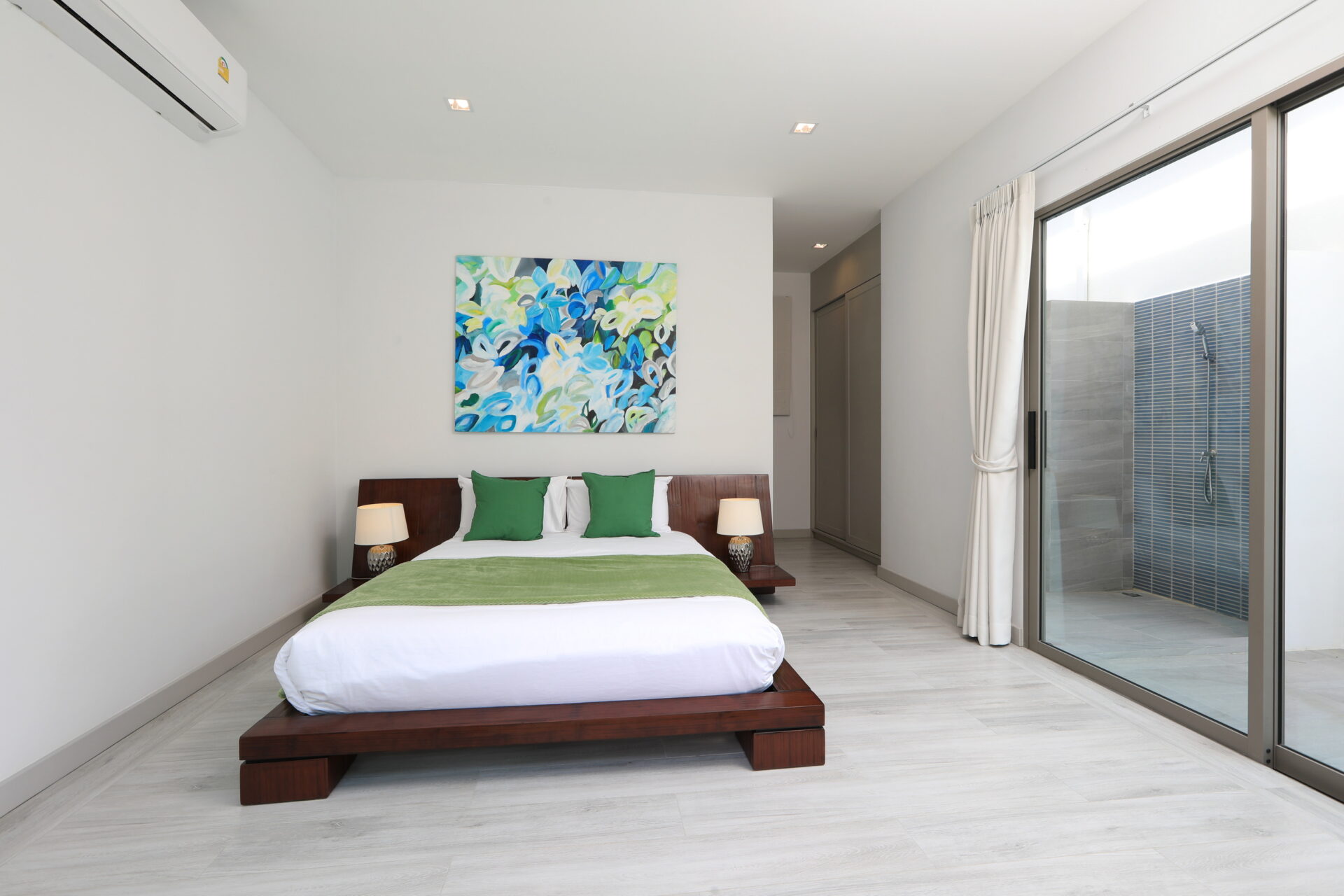 Modern bedroom with a low wooden platform bed, white linens, green pillows, a colorful abstract painting, and a glass door opening to an outdoor shower area.