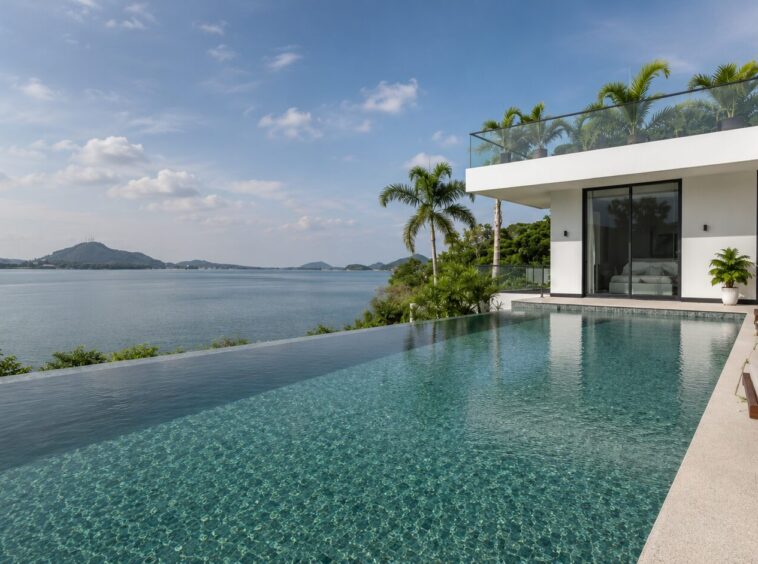 Infinity-edge pool beside a modern white villa, overlooking a calm sea and distant hills with palm trees nearby.