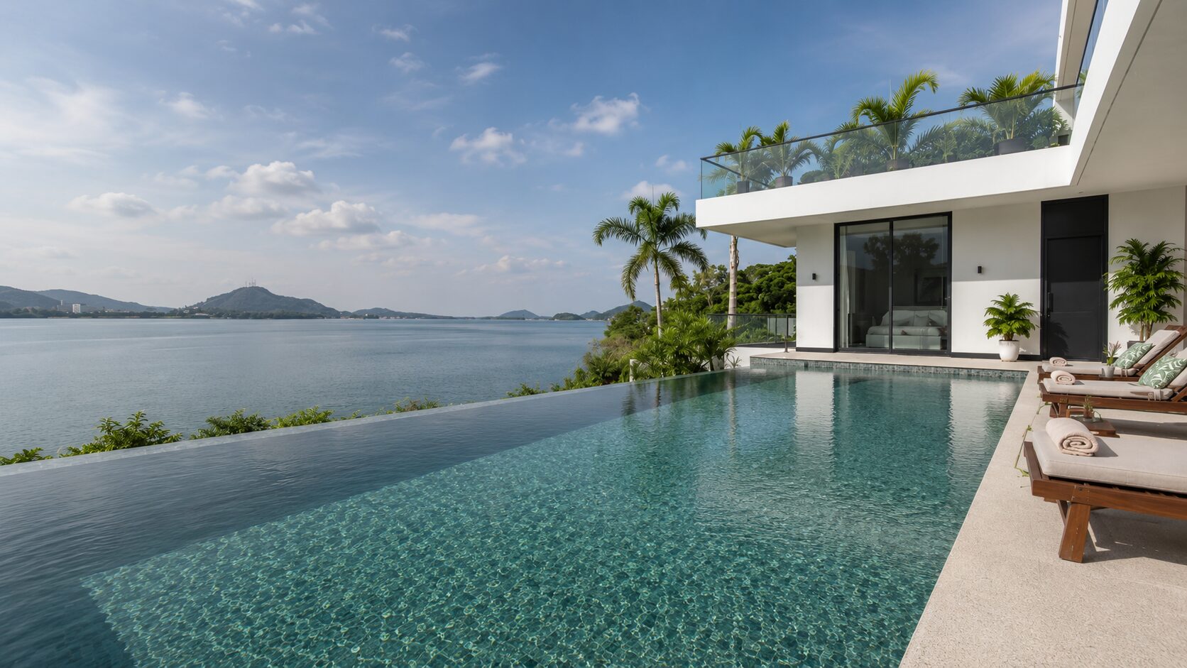Infinity-edge pool beside a modern white villa, overlooking a calm sea and distant hills with palm trees nearby.
