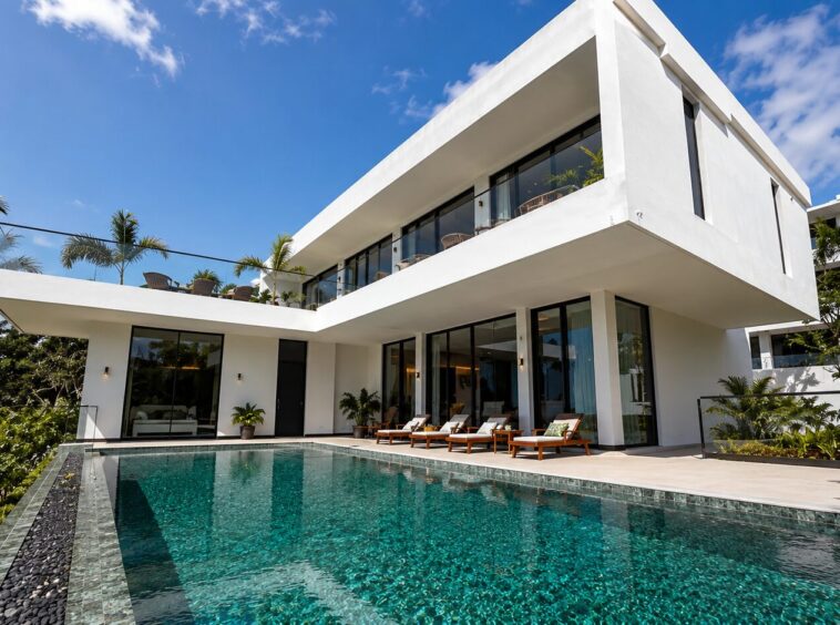 Modern white villa with glass walls, a rectangular pool, and palm trees under a bright blue sky; outdoor lounge chairs line the pool deck.