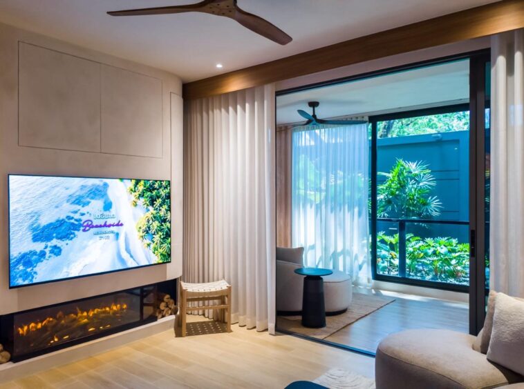 Modern living room with a wall-mounted flat-screen TV above a linear gas fireplace, cream-curtain-clad windows, and exposed wooden details.