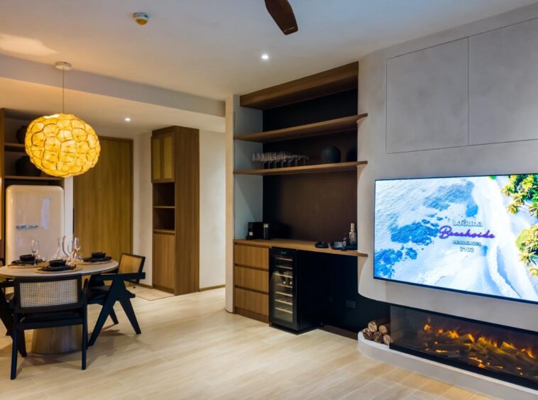 Open-plan living area with a round dining table, wooden chairs, and a warm pendant light near a compact kitchen and fridge.