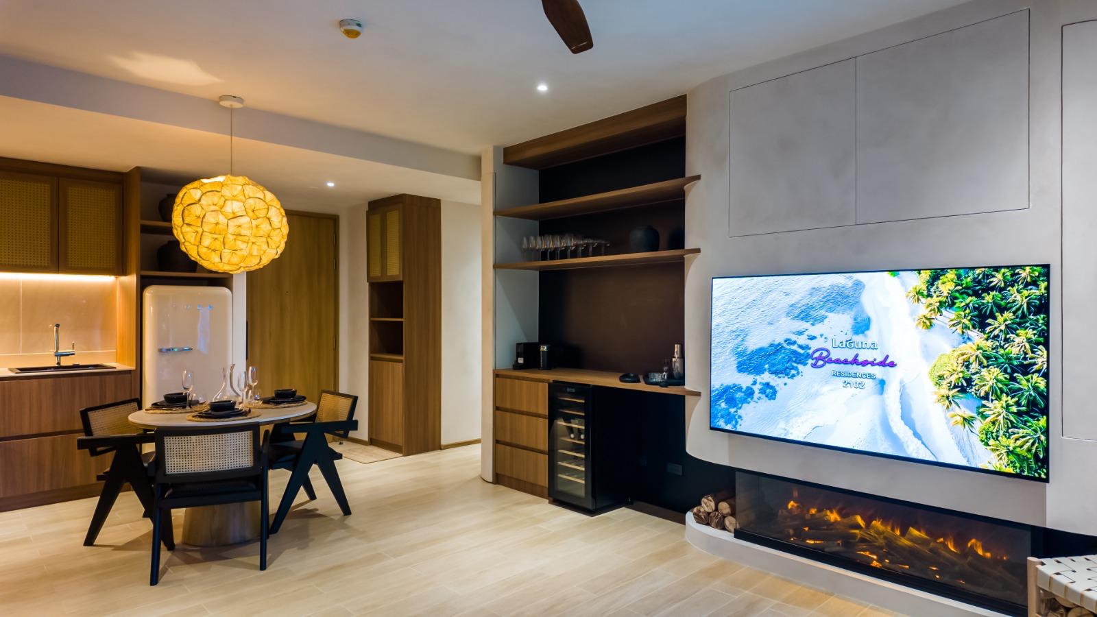 Open-plan living area with a round dining table, wooden chairs, and a warm pendant light near a compact kitchen and fridge.
