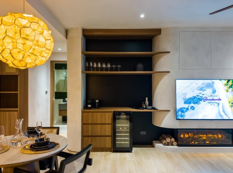 Contemporary dining and living area with a round table set for four, a large yellow pendant lamp, and a built-in entertainment wall with a fireplace and TV.