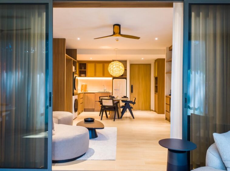 Open-plan living space viewed through sliding glass doors: kitchen with wooden cabinets, round dining table, woven pendant light, and ceiling fan.