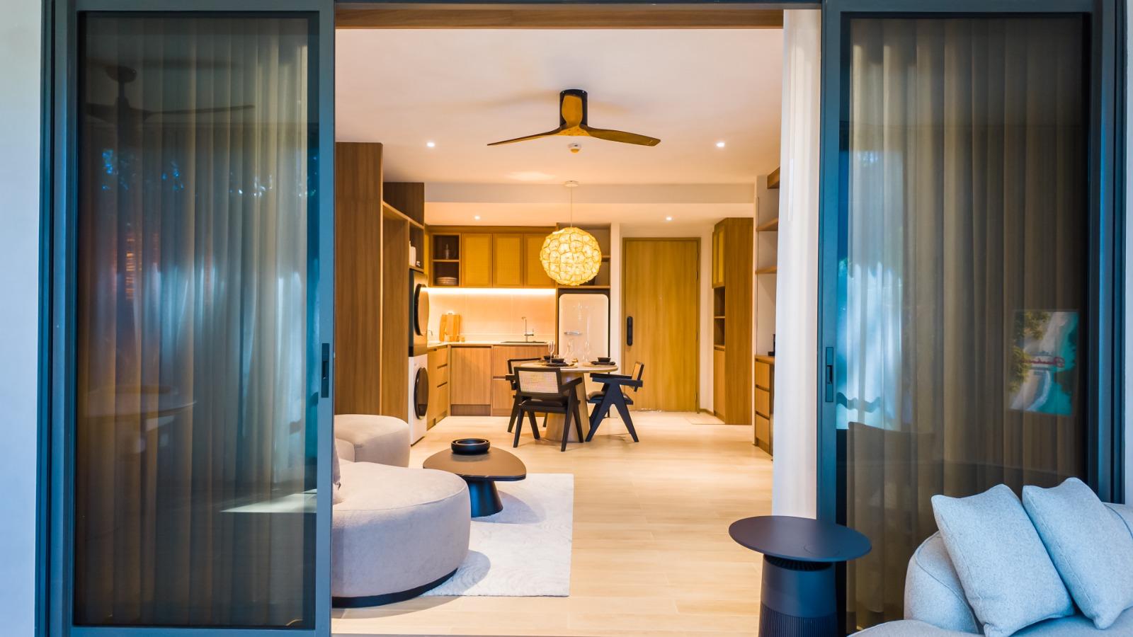 Open-plan living space viewed through sliding glass doors: kitchen with wooden cabinets, round dining table, woven pendant light, and ceiling fan.