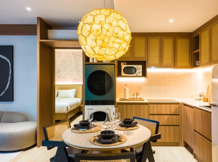 Open-plan kitchen and dining area with a round table for four under a large yellow pendant light; laundry stack, microwave, and white SMEG fridge visible in warm wood cabinetry.