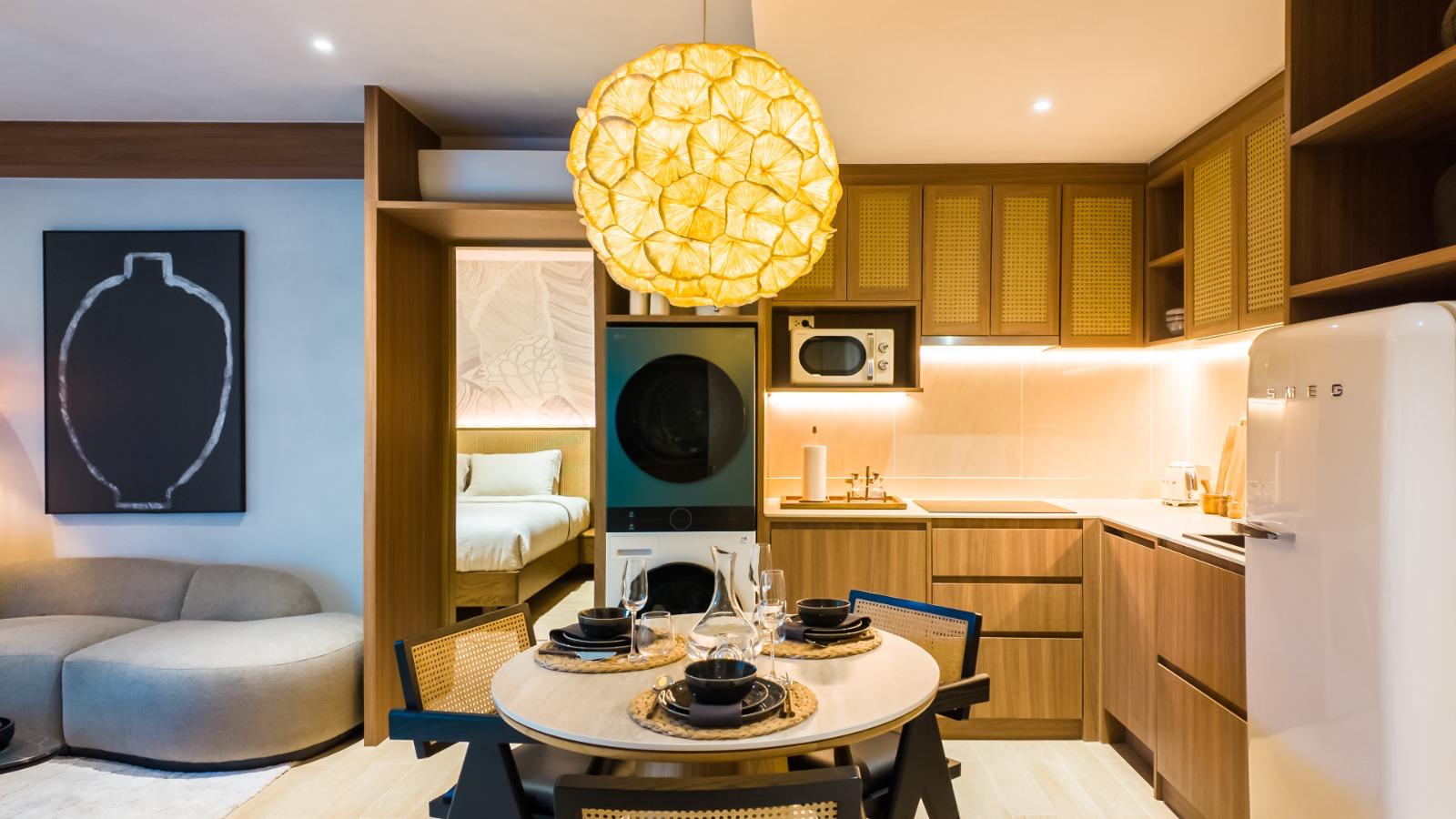 Open-plan kitchen and dining area with a round table for four under a large yellow pendant light; laundry stack, microwave, and white SMEG fridge visible in warm wood cabinetry.