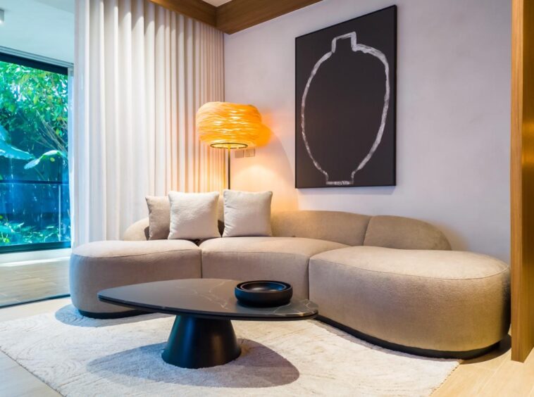 Modern hotel suite living area: beige curved sofa, black round coffee table, warm floor lamp, large window with greenery, and abstract wall art.