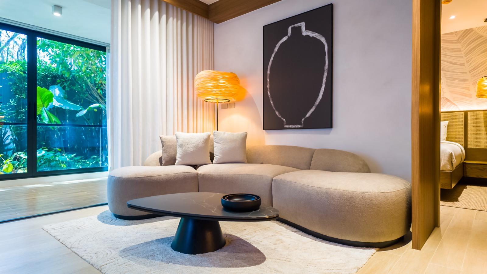 Modern hotel suite living area: beige curved sofa, black round coffee table, warm floor lamp, large window with greenery, and abstract wall art.