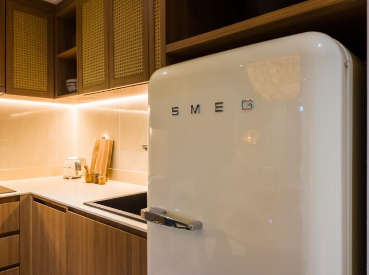 Cream SMEG refrigerator with chrome handle in a warm, wood-toned kitchen under recessed lighting.