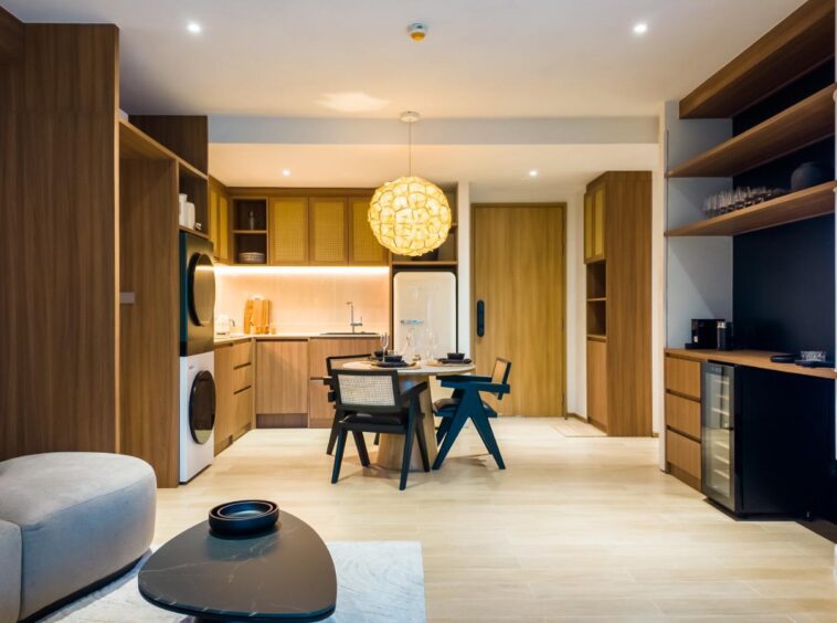 Open-concept living area with wooden kitchen cabinets, round woven pendant light, and a dining table at center.