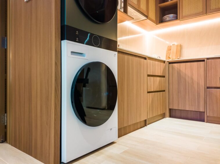 Front-loading washing machine under a stacked dryer in a wood-furnished kitchen with warm under-cabinet lighting.