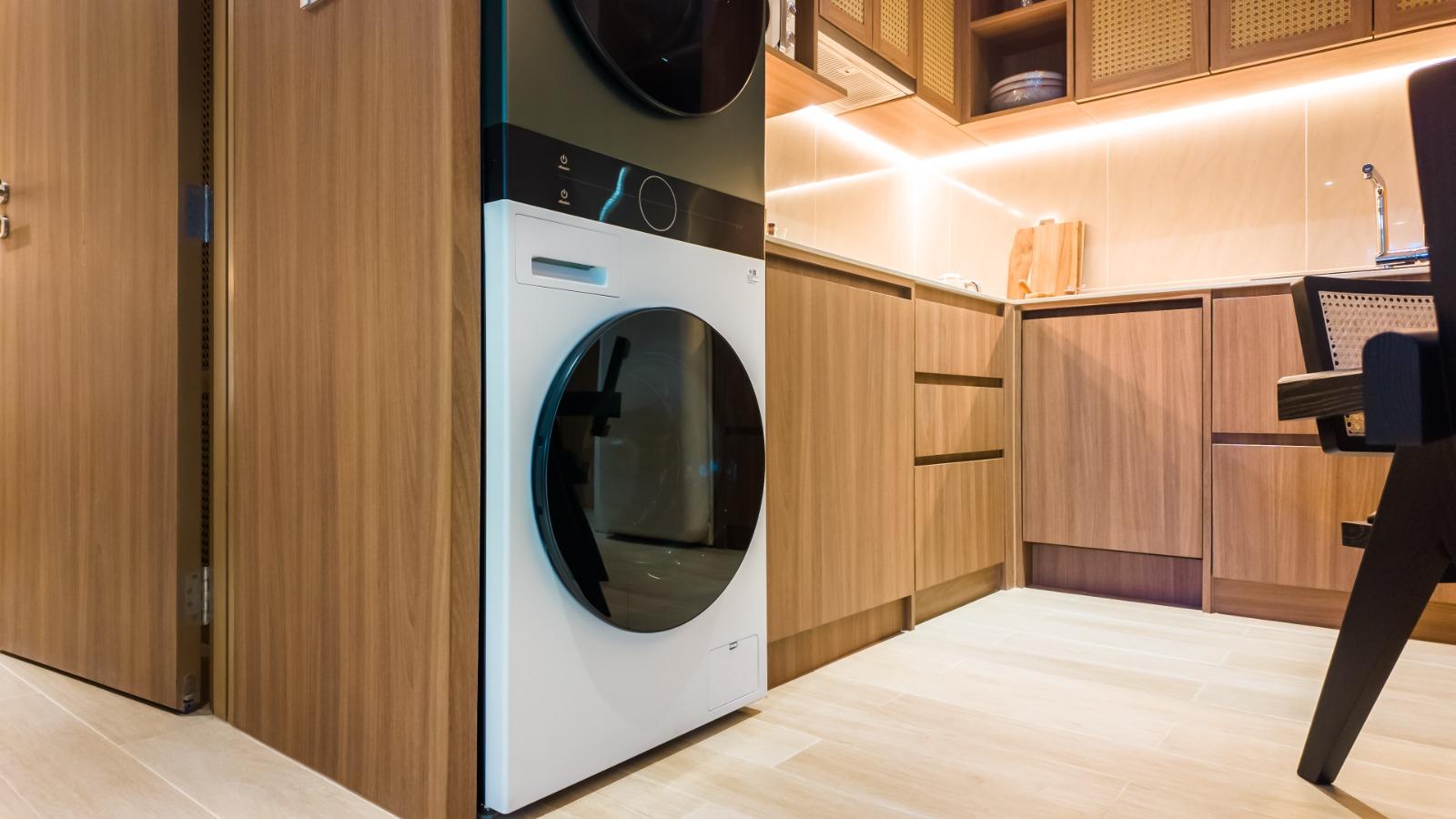 Front-loading washing machine under a stacked dryer in a wood-furnished kitchen with warm under-cabinet lighting.