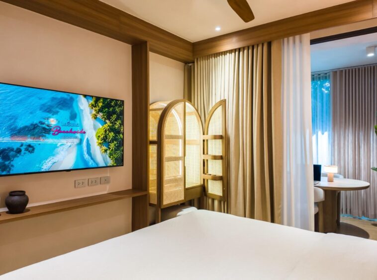 Hotel room interior: wall-mounted flat-screen TV, wooden shelf with decorative pots, and a woven folding room divider by beige curtains.