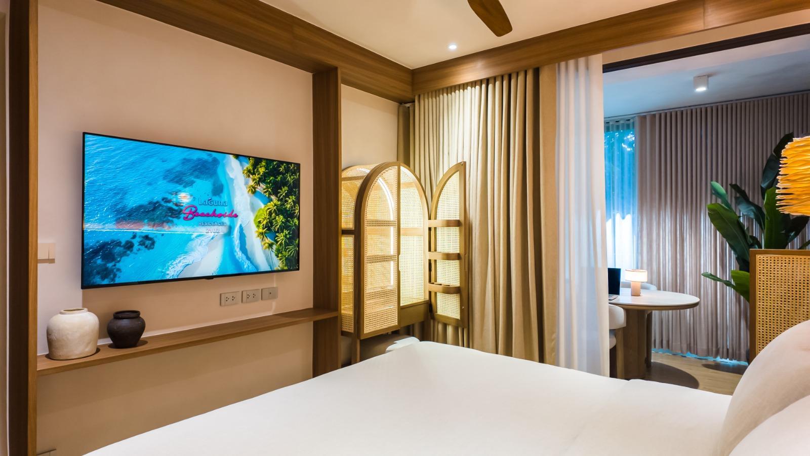Hotel room interior: wall-mounted flat-screen TV, wooden shelf with decorative pots, and a woven folding room divider by beige curtains.