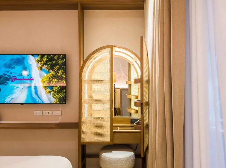 Hotel room: wall TV playing a beach scene, a wicker-panel vanity with a circular light mirror, and beige curtains.