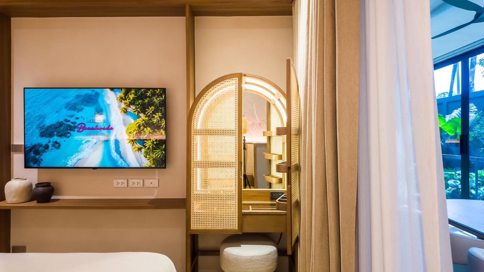 Hotel room: wall TV playing a beach scene, a wicker-panel vanity with a circular light mirror, and beige curtains.
