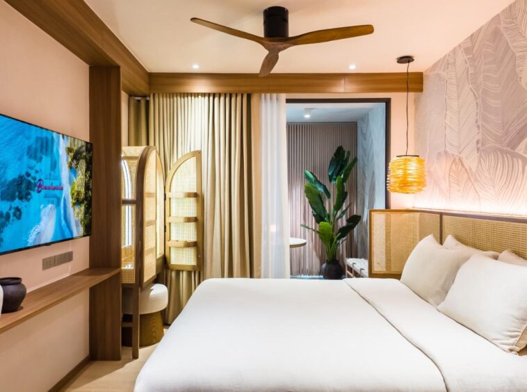 Large hotel room with a white-linen bed, woven headboard, and a wall mural; TV mounted on a wooden console to the left.
