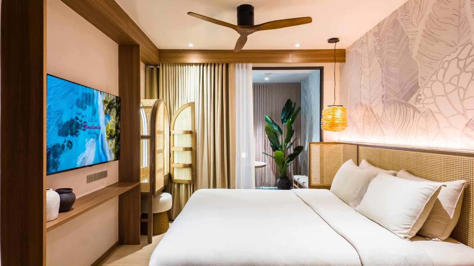 Large hotel room with a white-linen bed, woven headboard, and a wall mural; TV mounted on a wooden console to the left.