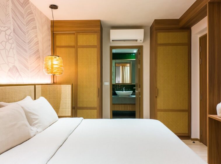 Hotel room interior with a white bed, woven headboard panels, and a warm hanging lamp on the left; wooden closets, doorway to an en-suite bathroom, and a wall-mounted TV on the right.