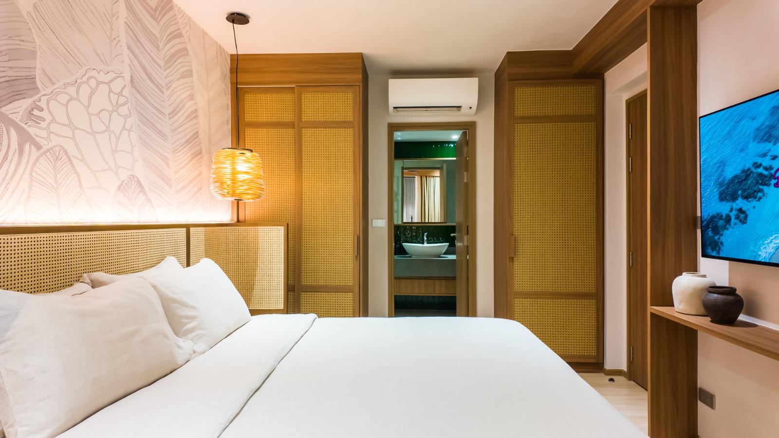 Hotel room interior with a white bed, woven headboard panels, and a warm hanging lamp on the left; wooden closets, doorway to an en-suite bathroom, and a wall-mounted TV on the right.