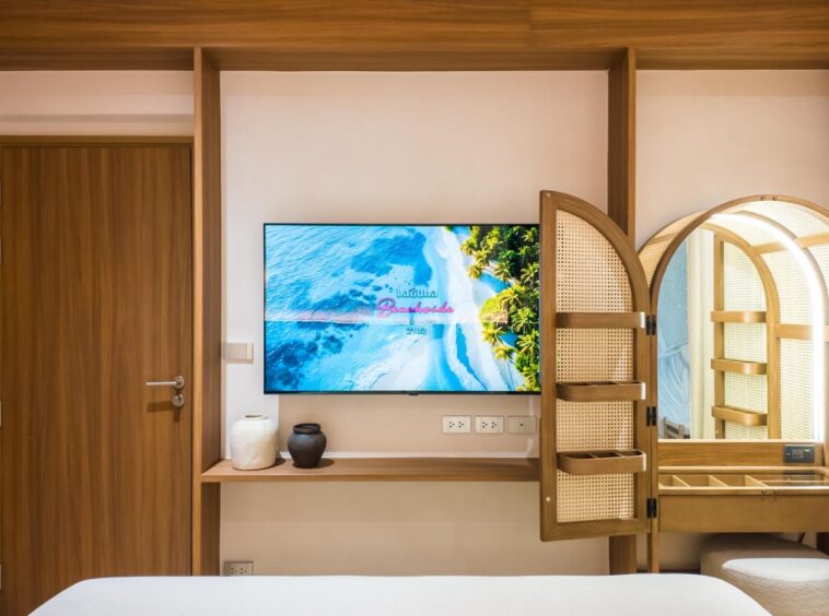 Hotel room interior with a wall-mounted TV opposite a bed, a wooden door, and a desk with decorative vases.