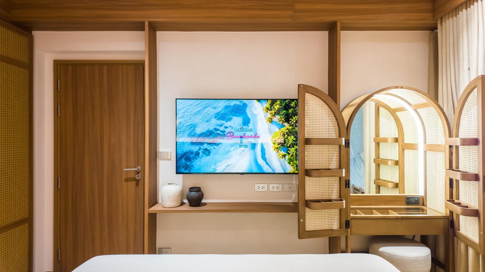 Hotel room interior with a wall-mounted TV opposite a bed, a wooden door, and a desk with decorative vases.