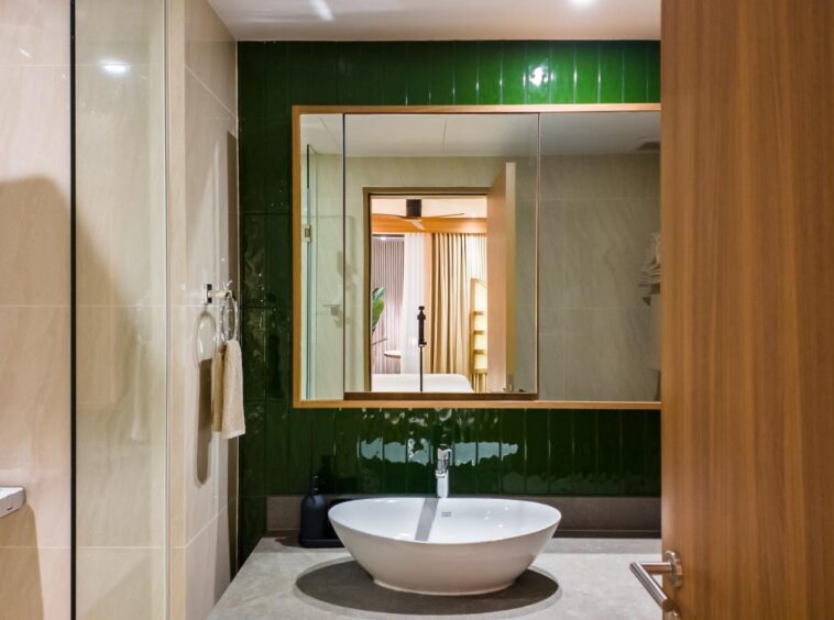 Modern bathroom with a round white vessel sink on a gray countertop, dark green glossy tiles behind a framed mirror; shower to the left and a wooden door to the right.
