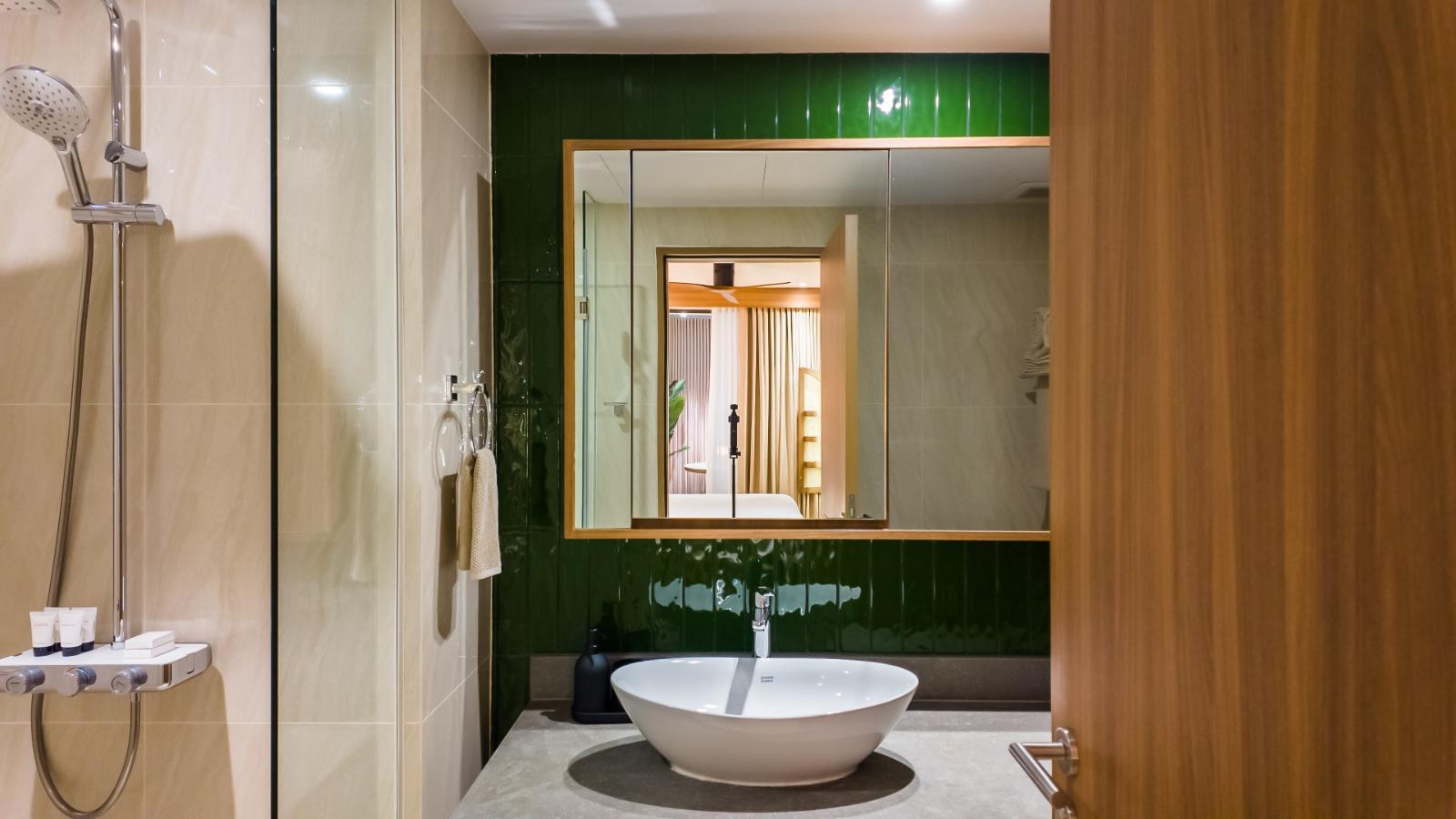 Modern bathroom with a round white vessel sink on a gray countertop, dark green glossy tiles behind a framed mirror; shower to the left and a wooden door to the right.