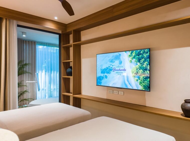 Twin hotel bedroom with two white beds, a wall-mounted TV, and built-in wooden shelves along the wall.