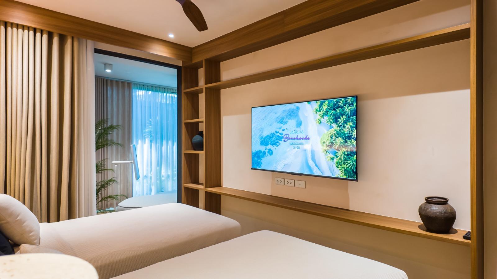 Twin hotel bedroom with two white beds, a wall-mounted TV, and built-in wooden shelves along the wall.