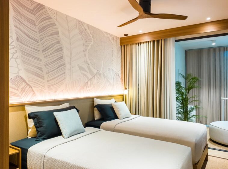 Twin beds with teal and white cushions in a hotel room; botanical wallpaper and warm wall lighting.