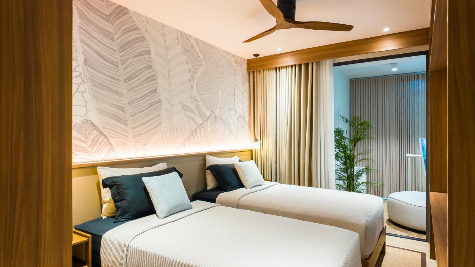 Twin beds with teal and white cushions in a hotel room; botanical wallpaper and warm wall lighting.