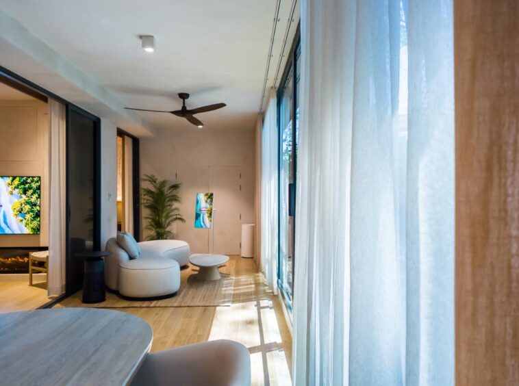 Bright modern living room with large floor-to-ceiling windows, sheer white curtains, and a curved white sofa with a coffee table in the center.