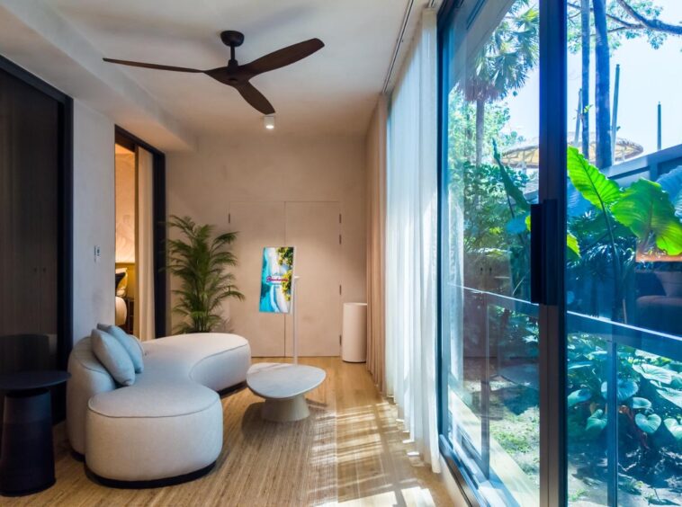 Modern living room with a curved white sofa, round coffee table, and a tall indoor plant facing floor-to-ceiling glass doors revealing a tropical garden outside. Ceiling fan and soft natural light create a bright, airy ambiance.