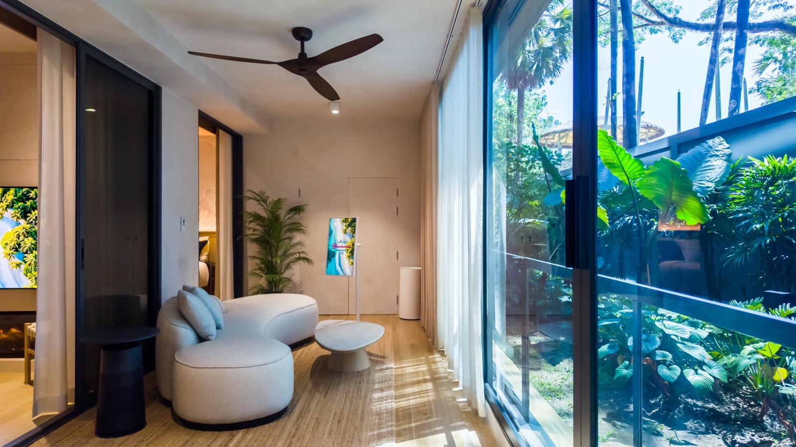 Modern living room with a curved white sofa, round coffee table, and a tall indoor plant facing floor-to-ceiling glass doors revealing a tropical garden outside. Ceiling fan and soft natural light create a bright, airy ambiance.