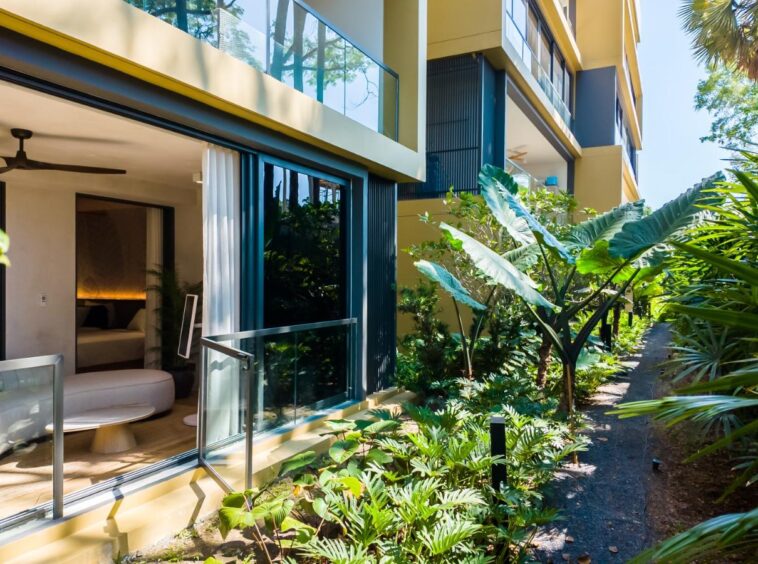 Ground-floor hospitality suite with glass walls open to a lush tropical garden path and exterior greenery outside the living area.